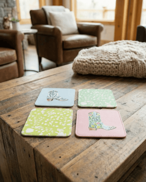Four rodeo cowboy boot print coasters with designs on a wooden surface in a cozy living room.