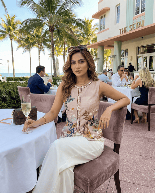 Woman sitting at an outdoor dining table with palm trees and a building in the background
